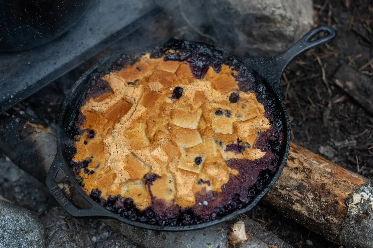 A cast-iron skillet of blueberry cobbler, biscuit topping browned, set on coals at the edge of a fire.