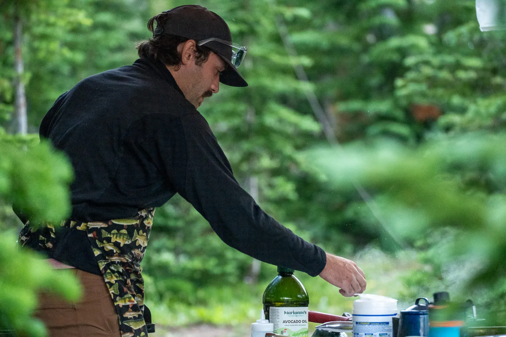 Nick in profile in a cap and sunglasses, working at an outdoor prep station, green forest behind.