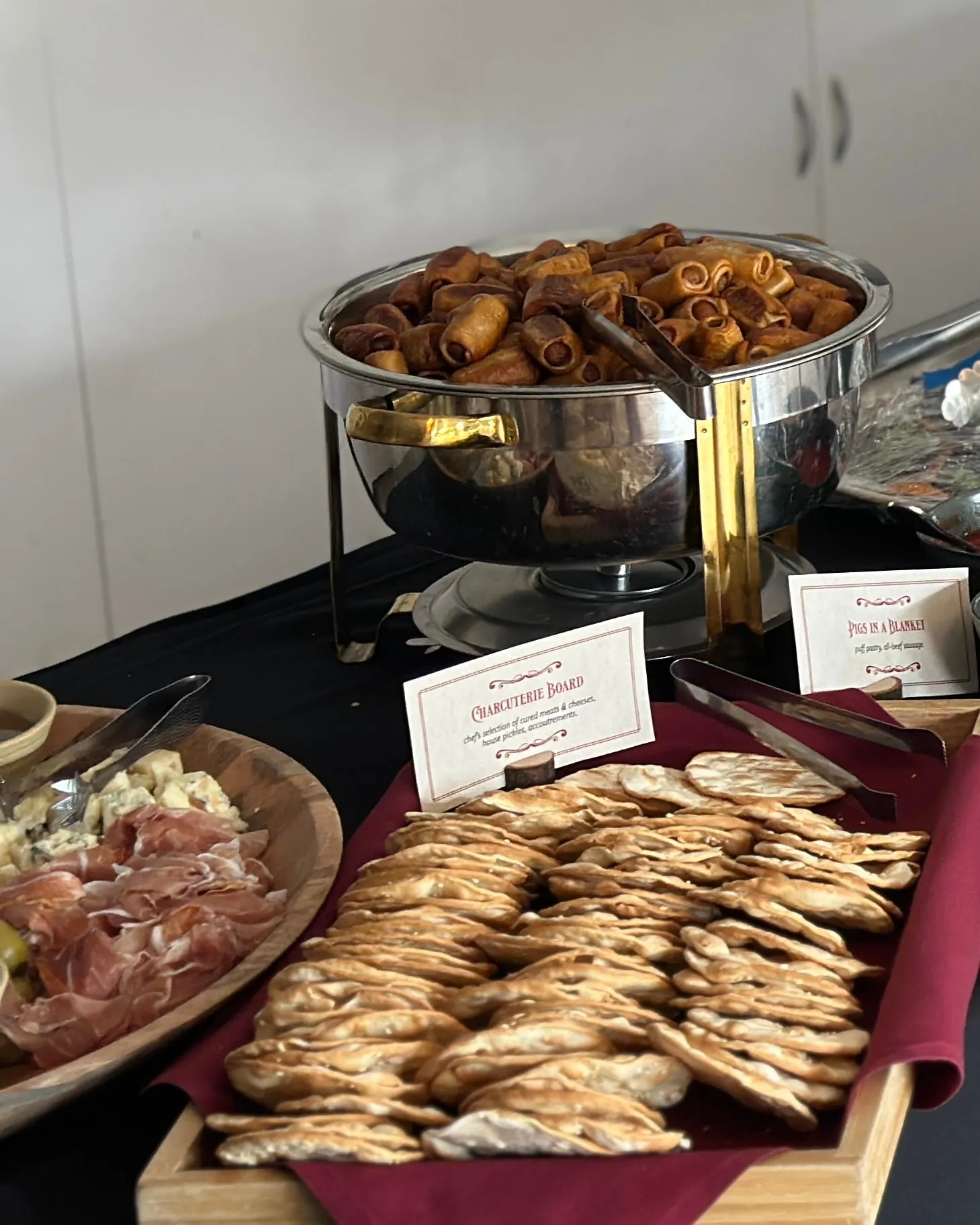 A catered spread on a black-draped table: a tray of golden pastries, a charcuterie board, and a chafing pan of hors d’oeuvres.