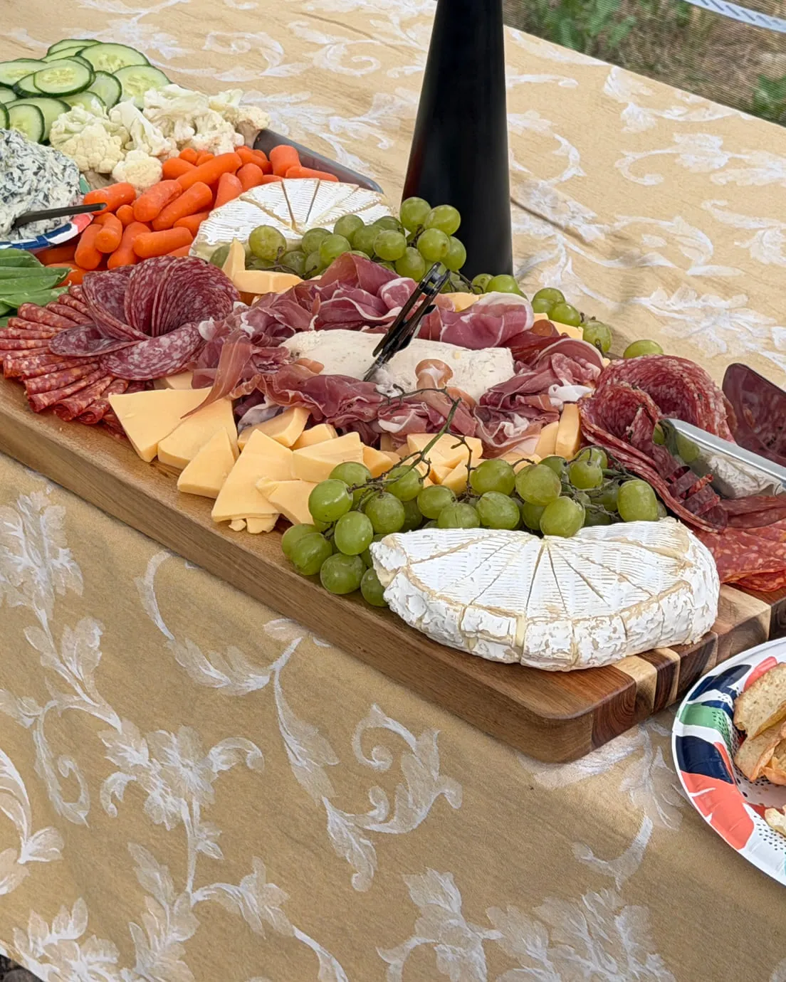 A grazing table set outdoors — cured meats, cheeses, grapes and crudités laid along a wooden board, pines behind.